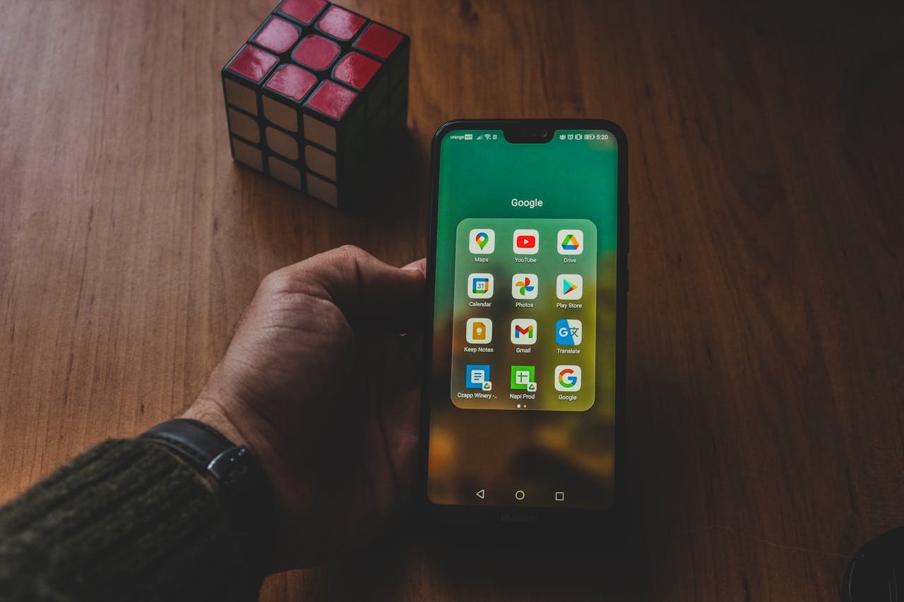 A hand holds a smartphone displaying various apps, next to a Rubiks cube on a wooden surface.